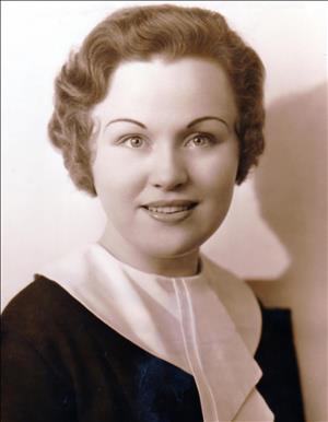 A portrait of a young white woman smiling at the camera. She has light brown hair and groomed eyebrows and is wearing a blouse with a large white collar